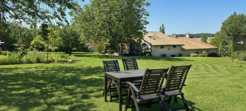 Tuin met houten tafel en stoelen, omgeven door gras en bomen onder een heldere blauwe lucht.