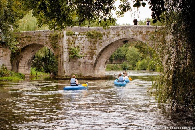 Twee kajakkers op een rivier onder een stenen brug, omgeven door groene bomen.
