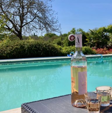 Fles rosé wijn en glazen naast een zwembad, omgeven door groen.